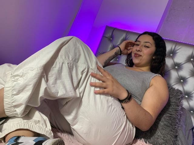 ScarlettParchMusa is laying on a bed, wearing a gray sleeveless top and white pants, with a relaxed smile, tattoo on her right arm, and purple room lighting.
