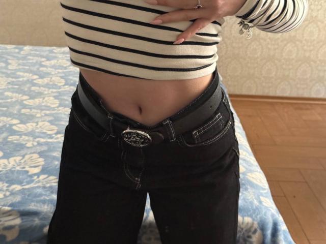 Siyana's striped shirt is lifted, exposing her flat stomach and black jeans with a gray belt, showing her navel. Her hand rests on her chest.