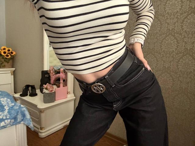 Siyana wearing a striped white and black top, dark jeans with a gray belt, and a silver buckle, hands in pockets, showing a slight belly and midriff.