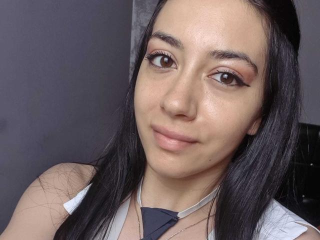 SerenaRin a la peau mate, les cheveux noirs, les yeux marron avec un trait d'eye-liner ailé, et un léger sourire. Elle porte un haut blanc sans manches avec une cravate noire..