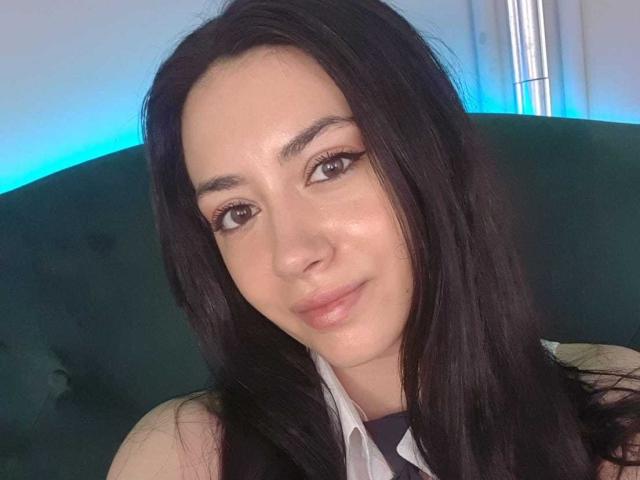 SerenaRin tem cabelos pretos longos, olhos castanhos e um sorriso discreto. Ela está vestindo uma camisa branca com uma gola preta, e seu rosto está levemente corado..