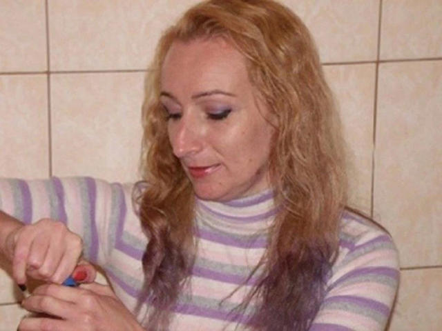 Pussyxxx, attraente con lunghi capelli biondi, indossa un maglione a collo alto a righe viola e bianco, tiene un piccolo oggetto rosso e blu nella mano destra, guarda in basso con un leggero sorriso..