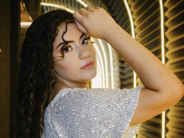 CaskiallX with curly dark hair, looking over shoulder, wearing a sparkly silver top, hand in hair, soft facial expression, warm light in background, medium skin tone, subtle makeup.