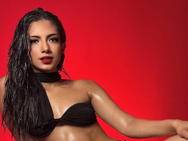 RousseRose a les cheveux noirs et humides, une peau brillante, et porte un haut noir à encolure dégagée qui couvre ses seins. Elle regarde l'appareil photo avec du rouge à lèvres rouge. Fond rouge..