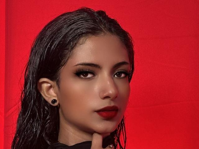 RousseRose has wet black hair, dark eyeliner, red lipstick, and black ear gauges. Her nose is small, and she's touching her chin with a slight smile.