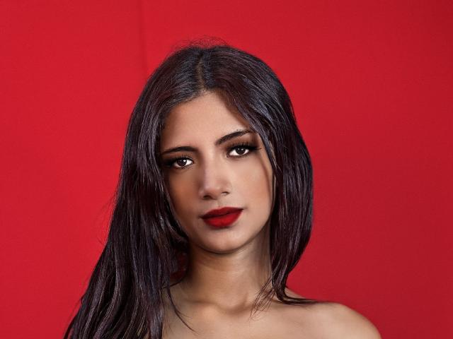 RousseRose a les cheveux noirs et lisses, du rouge à lèvres, et des yeux marrons. Elle regarde directement l'appareil photo avec une expression neutre. Elle est sur un fond rouge uni..