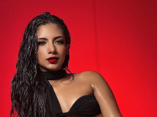 RousseRose con i capelli neri bagnati, rossetto rosso e un top nero a halter, che mostra il seno e un'espressione seria, su uno sfondo rosso brillante..