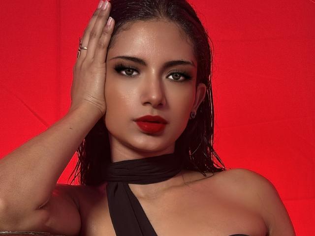 RousseRose hat feuchtes, braunes Haar, roten Lippenstift und dunkle Make-up. Sie trägt ein schwarzes Top mit Halter und berührt mit einer Hand, an der ein Ring zu sehen ist, ihren Kopf. Roter Hintergrund..