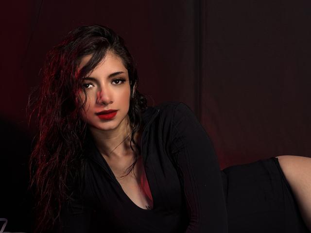RousseRose with curly black hair, red lipstick, and black top, lying down, showing a side of her bare ass, dark red light on her face, and serious expression.