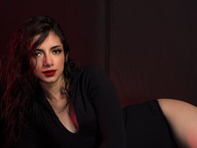 RousseRose, con capelli scuri, ricci e umidi, rossetto rosso e vestito nero, sdraiata, che mostra il sedere, guarda verso la telecamera, sfondo scuro con luce rossa..