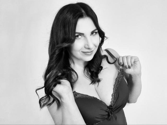 SandraSmile with dark, wavy hair, smiling, wearing a black lace bra, pulling straps, showing her tits, and holding her bra strap, in a black-and-white photo.