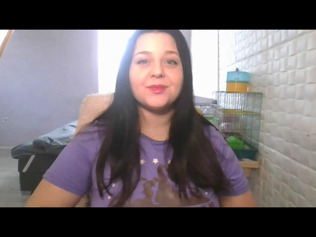 EricaLaLovely mit langem, schwarzem Haar, einem lila Hemd, kleinen Brüsten und einem leichten Lächeln, sitzt in einem Raum mit einem Vogelkäfig und einer weißen Ziegelwand..