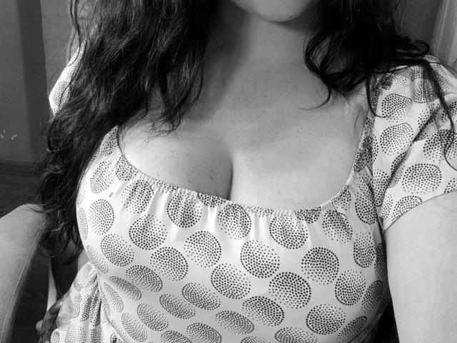 black-and-white photo of EricaLaLovely with wavy dark hair, wearing a patterned dress, showcasing her big, round, soft boobs and visible cleavage, sitting in a chair.