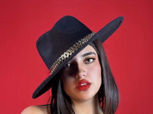 Abrilossa with black hat, gold chain, and red lipstick, looking straight at camera, has dark brown hair, and a serious expression, against a red background.