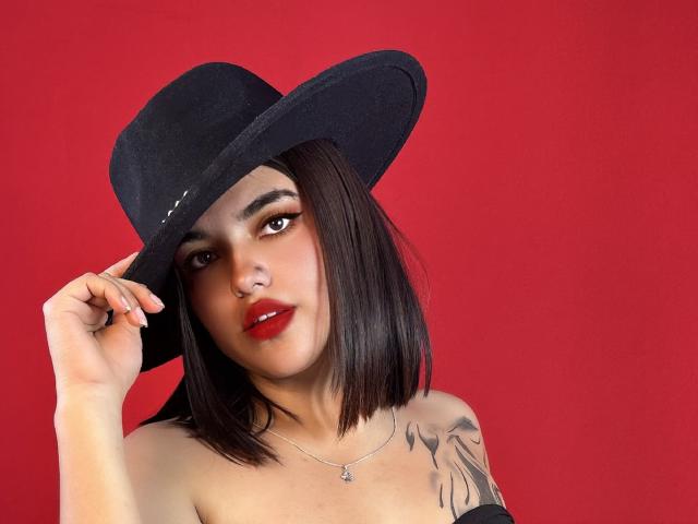 Abrilossa with dark bob, wearing a black hat, red lipstick, necklace, and tattoo on shoulder, holding hat against red background, showing off her tits and cute face.
