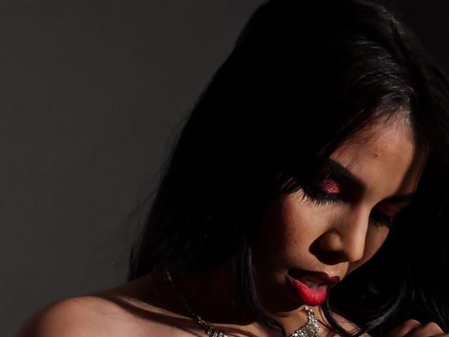 EmmaLasso with dark hair, red lipstick, and eye makeup, looking down, showing her ass, wearing a necklace, in low light, shadowy background, her face partially in shadow.