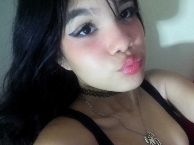 MidnightHarpy with black hair, blue eyes, and pink lips, pouting. She's wearing a black choker, necklace, and a black top, showing her tits. Her face is slightly flushed.