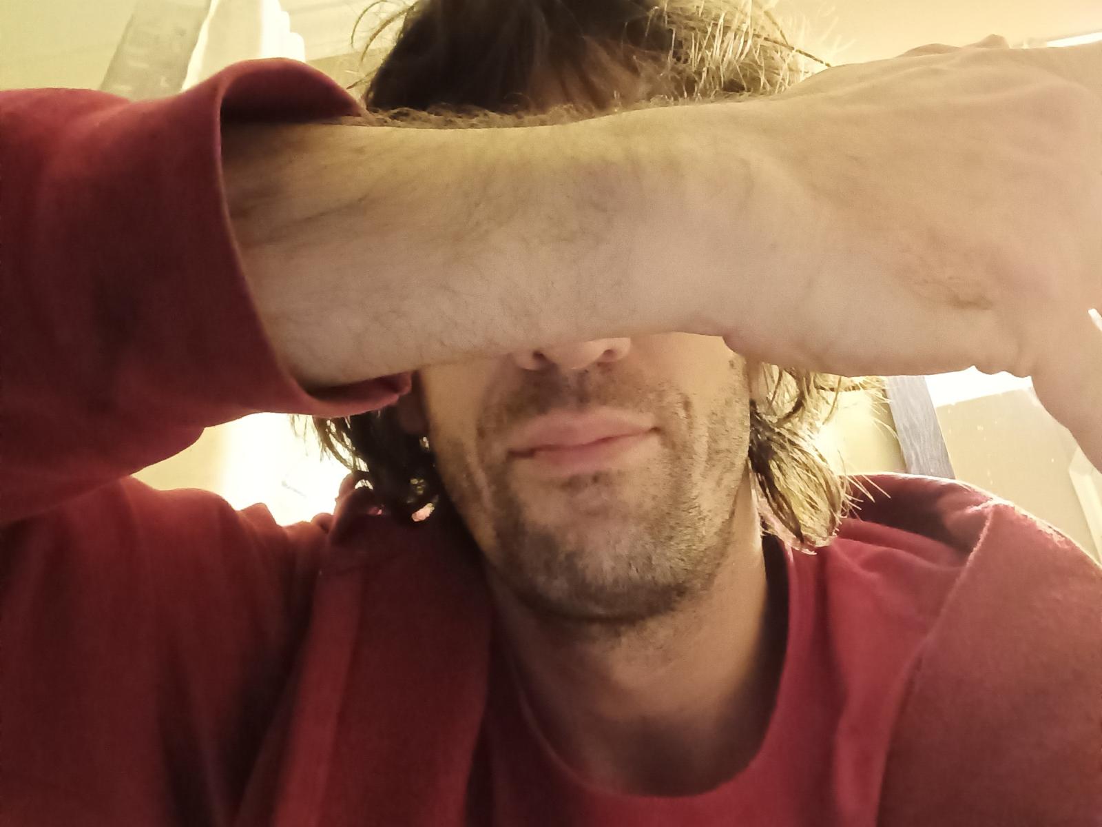 EmaBoyluvX with messy brown hair, slight beard, and red shirt, covering eyes with a hand, smiling slightly, visible nose and mouth. EmaBoyluvX's hand has visible veins and hair.
