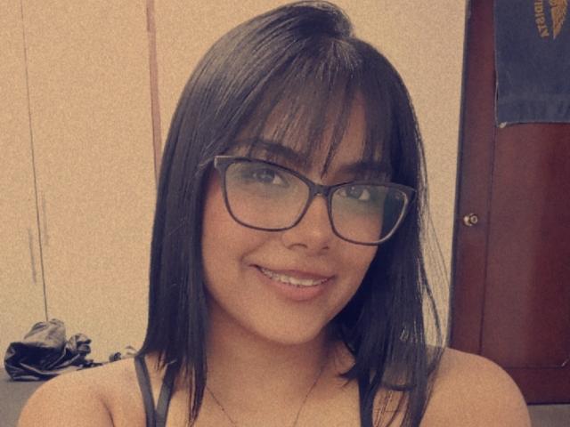 EmilyRobeerts has black, straight hair, glasses, and is smiling with her teeth showing. She's wearing a gray tank top, and her tits are slightly visible.