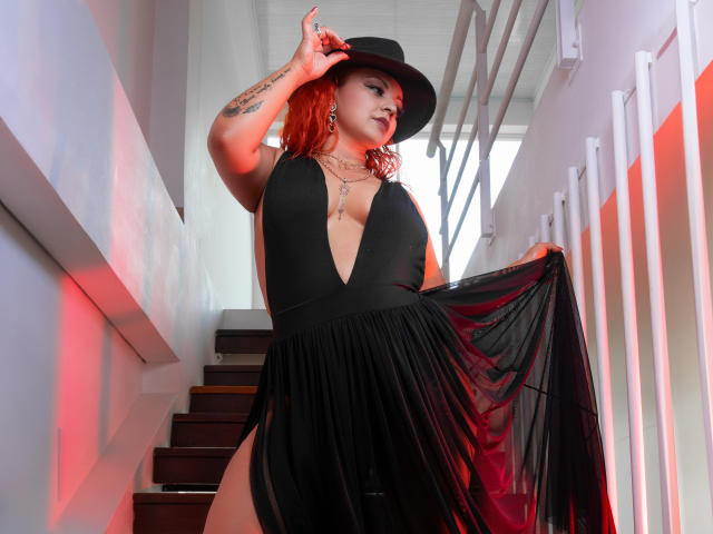LillyBigAss with red hair, black deep V dress, sheer black skirt, black hat, holding skirt up, showing big tits, tattoos on arm, stairs, red and white lighting.