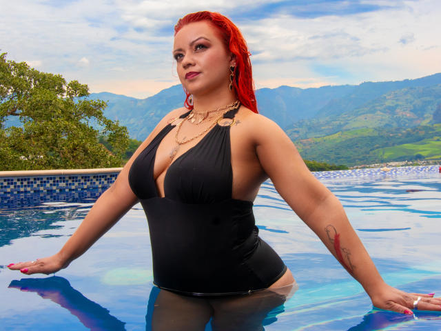LillyBigAss with red wet hair, black halter one-piece, large tits, tattoo on right arm, standing in blue-tiled pool, looking at camera, mountains and green trees in background.