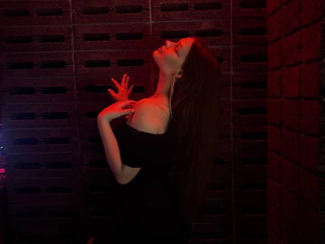 SirenScarlette with long brown hair, off-shoulder black dress, red light, eyes closed, hand on chest, hand on wall, standing against dark brick, sensual pose, delicate earrings.
