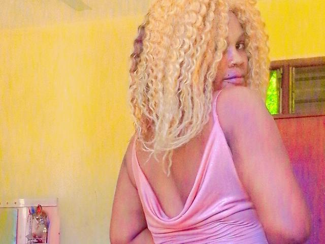 Pepitaa with blonde curly hair, pink tank top, showing her ass, looking over her shoulder, nose ring, yellow wall, and a window in the background.