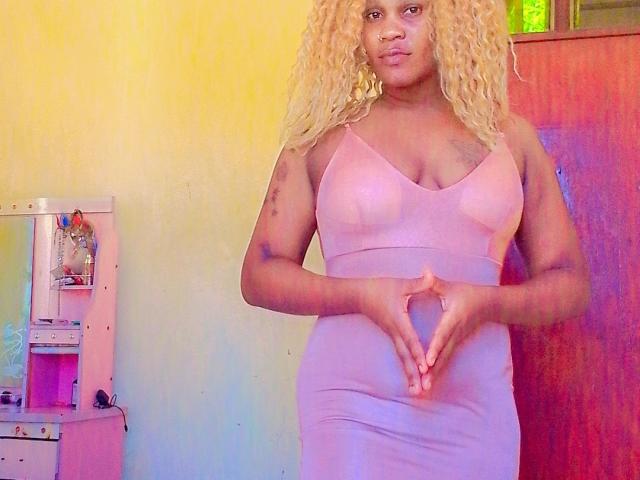 Pepitaa with blonde curly hair, wearing a pink, tight, cleavage-revealing dress, making a heart shape with her hands, standing in a room with a wooden door.