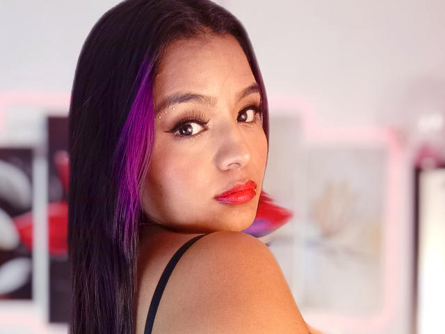SofiLoveTorez has dark black hair with purple highlights, wearing red lipstick, black strap top, and looking over her shoulder with a serious expression. Her tits are partially visible.