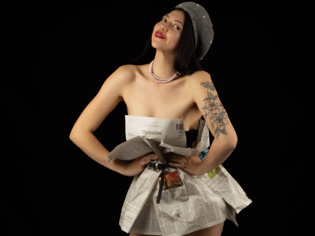 MissAuroraa's tits are exposed, wearing a gray hat and pink necklace, holding crumpled newspaper, tattoo on right arm, against black background, looking up with red lips.
