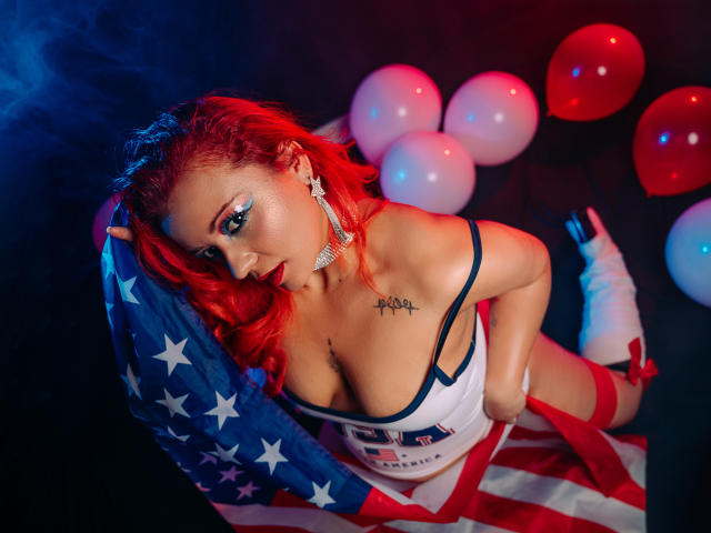 LillyBigAss with red hair, wearing a star-spangled top, holding a flag, and white boots, has big tits, a tattoo on her chest, and red balloons in the background.