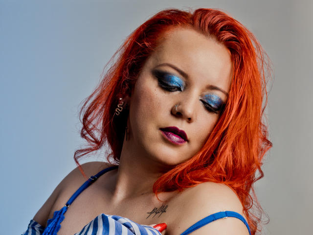 LillyBigAss with bright red hair, blue eyeshadow, purple lipstick, and a blue top, showing a tattoo on her right chest, looking down with a slightly melancholic expression.