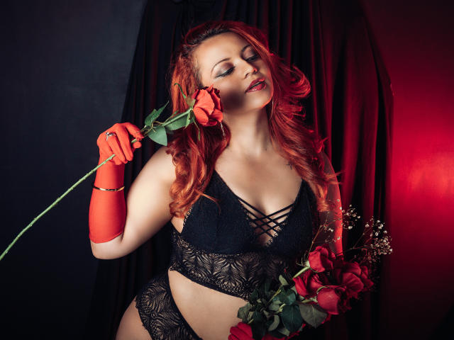 LillyBigAss with red hair, black lace bra, and panties, holding a rose, wearing red gloves, eyes closed, sexy, red and black lighting, red roses in hand.