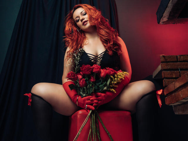 LillyBigAss with red hair, black lace dress, and red gloves, sitting with legs spread, holding red roses and greenery, on red stool, wearing black stockings.
