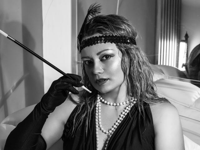 Black-and-white photo of LillyBigAss with wet, wavy hair, wearing a headband with a feather, black gloves, pearl necklace, holding a cane, looking seductively at the camera.