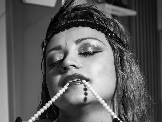 Black-and-white photo of LillyBigAss with wet, messy hair, eyes closed, pearl necklace in mouth, shiny eyeshadow, nose piercing, and black headband. LillyBigAss's lips are slightly parted.