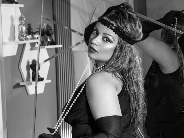 Black-and-white photo of LillyBigAss with wavy hair, wearing a black dress, gloves, pearl necklace, and headpiece, holding a fan, looking over her shoulder with a seductive expression.