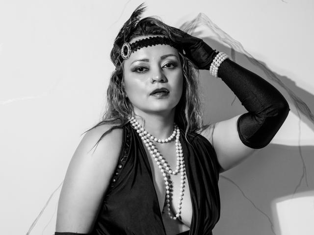 Black-and-white photo of LillyBigAss with wavy hair, wearing a headpiece, pearl necklace, and long black gloves, hand in hair, showing her tits, serious expression.