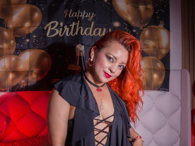 LillyBigAss with bright red hair, black lace-up dress, red lipstick, and earrings, sitting on red and white quilted couch, smiling at camera, looking sexy and confident.