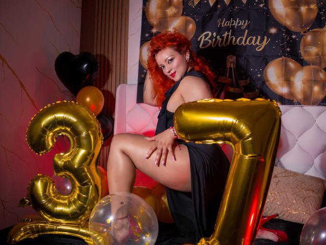 LillyBigAss with red hair, wearing a black dress, sitting on a bed with golden "3" balloons, showing her ass, surrounded by balloons and a plush toy.