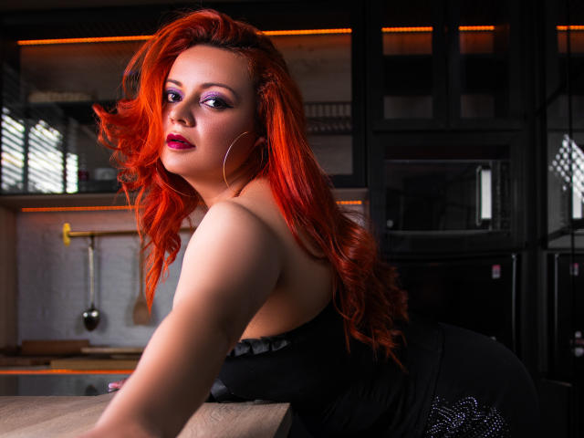 LillyBigAss with red hair, purple eyeshadow, and red lipstick, leaning on a kitchen counter, wearing a black dress, showing her ass, with hoop earrings, in a dark room.
