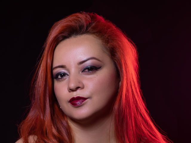 LillyBigAss has red hair, fair skin, dark eyeliner, and shiny red lipstick, with a slight smile and soft light highlighting her face. She's looking slightly to the left.