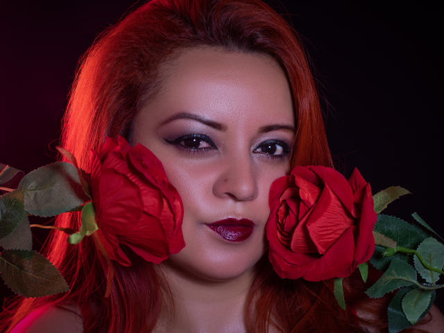 LillyBigAss with red roses on her cheeks, glossy dark red lipstick, and subtle eyeliner, has red hair and a serious expression, against a dark background.