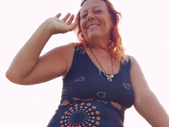 BarbaraBowie has red hair, smiling, wearing a black dress with orange patterns, and a necklace with a butterfly pendant, waving with her right hand.