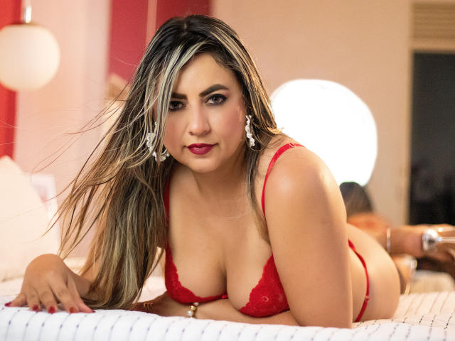 AmandaLeon, lying on a bed, wearing red lace lingerie, showing her tits, with long blonde and black hair, and red lipstick, looking at the camera with a slight smile.