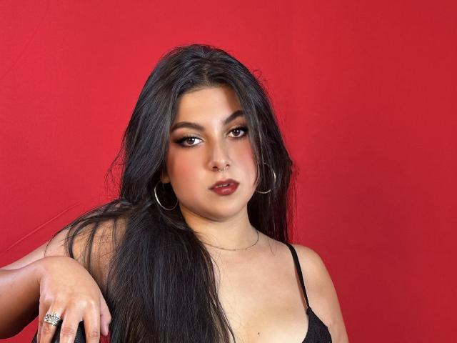 SamanthaOssa ha lunghi capelli neri, occhi marroni e rossetto scuro. Indossa un reggiseno nero, orecchini a cerchio e un anello. La sua mano è appoggiata sul ginocchio. Sfondo rosso..