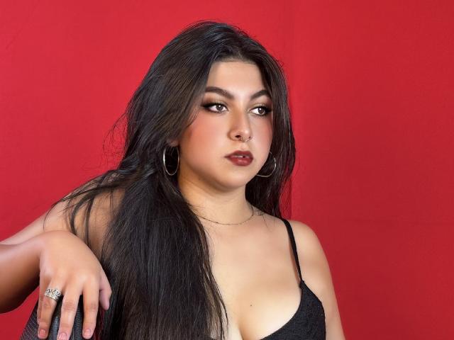 SamanthaOssa mit langem, schwarzem Haar, die ein schwarzes Top mit Spaghetti-Trägern, Creolen und einen Ring trägt, vor einem roten Hintergrund, wobei ihre Brüste und ein ernster Gesichtsausdruck zu sehen sind..