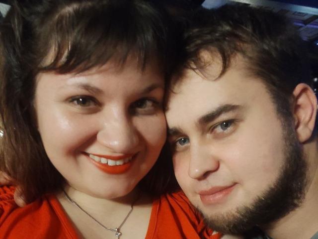 AlisaNAleks with orange lipstick, smiling, has chubby cheeks, and is next to a bearded guy with blue eyes, both have fair skin and dark brown hair.