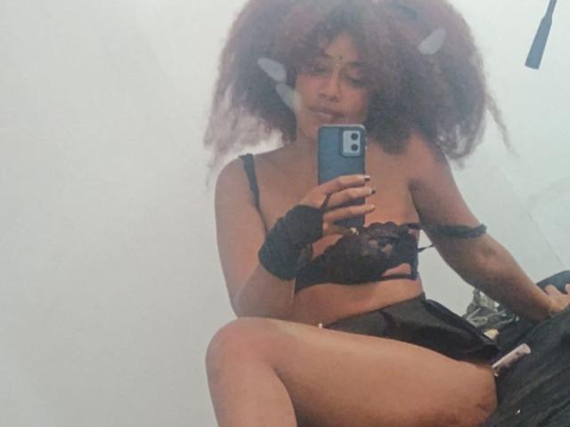 DalilaHeinz with curly afro, black lace bra, and fingerless gloves, sitting, holding phone, black shorts, showing tits, looking at mirror, in a slightly foggy room.