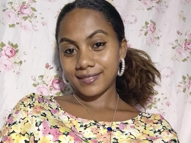 KennyReah has dark brown skin, brown eyes, and a slight smile. She's wearing a floral top, pearl earrings, and a necklace with a blue gem. Her ponytail is back.